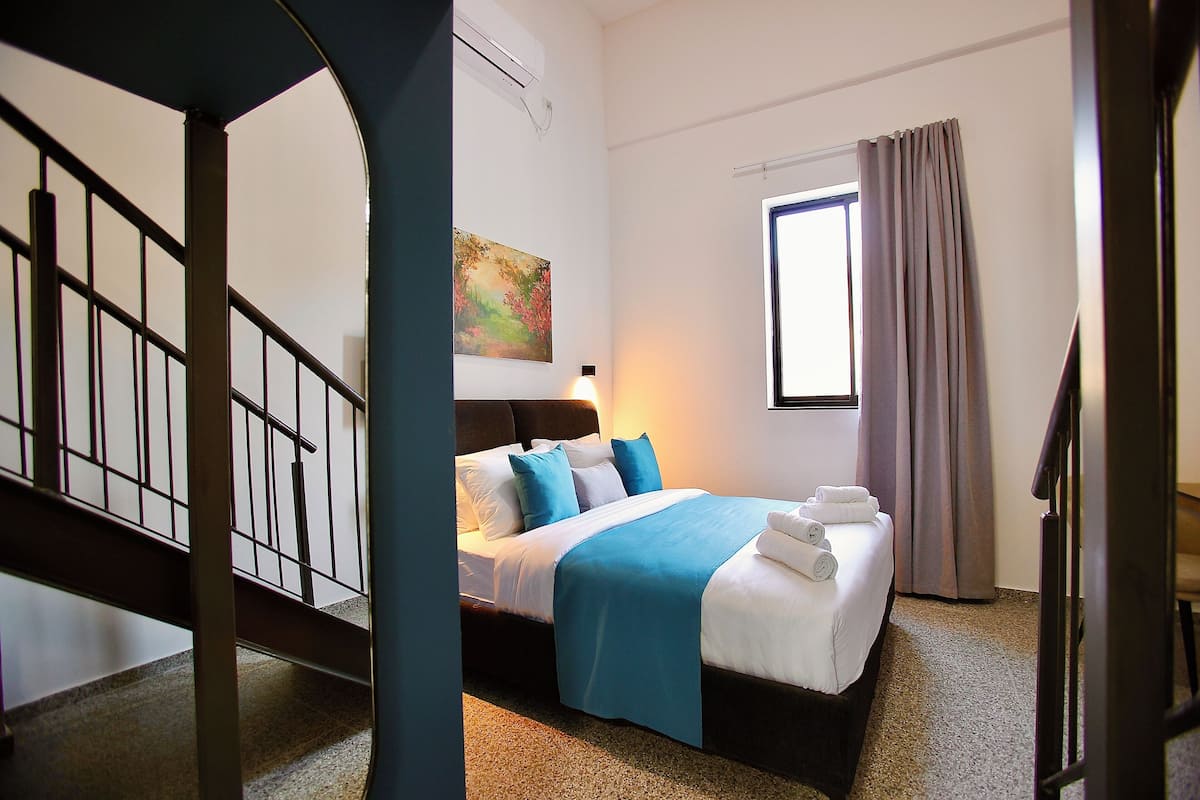 Una habitación de hotel con cama, un cuadro en la pared, una ventana con cortinas y una escalera.
