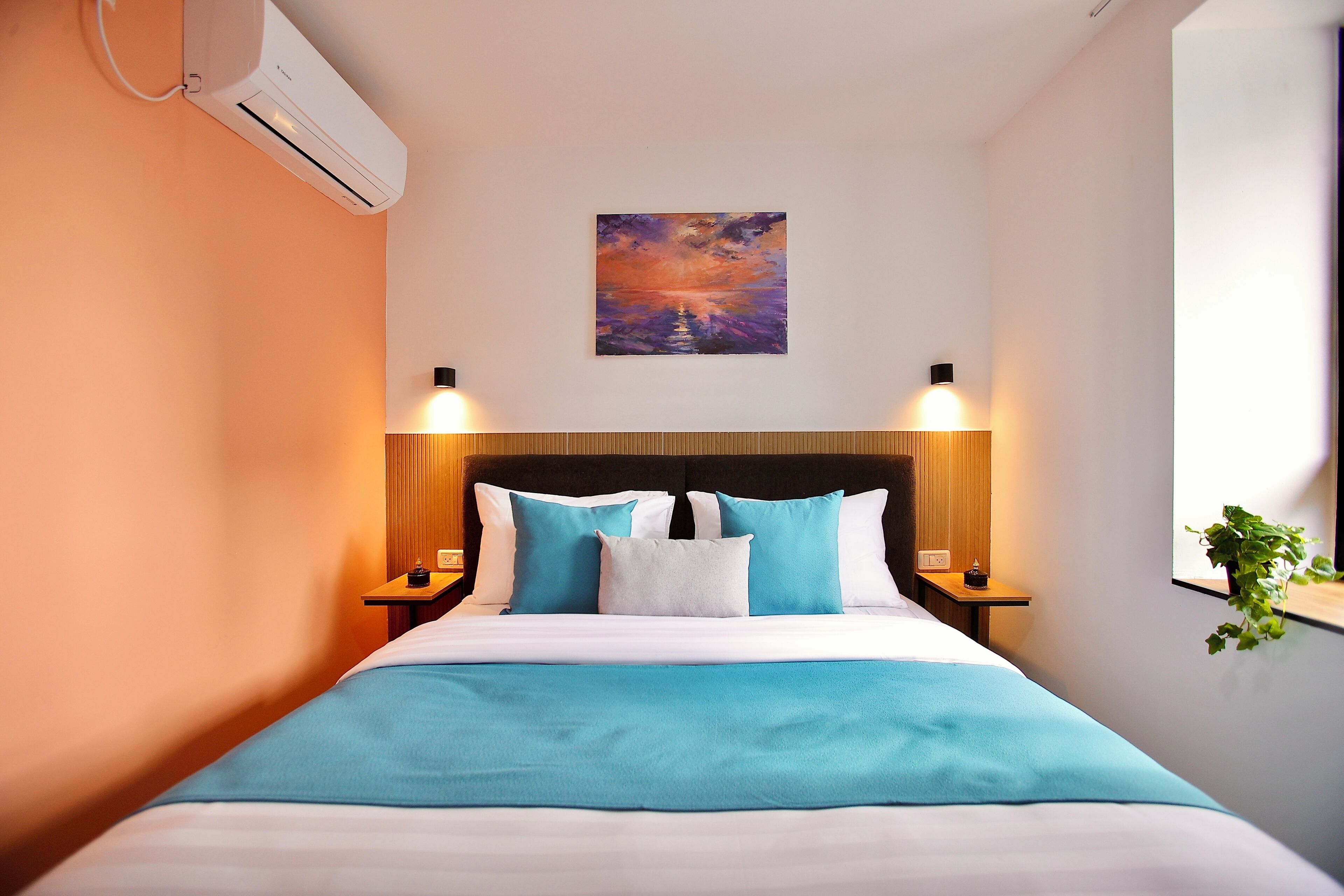 A hotel room with a bed, a painting, two bedside lamps, and an air conditioning unit.