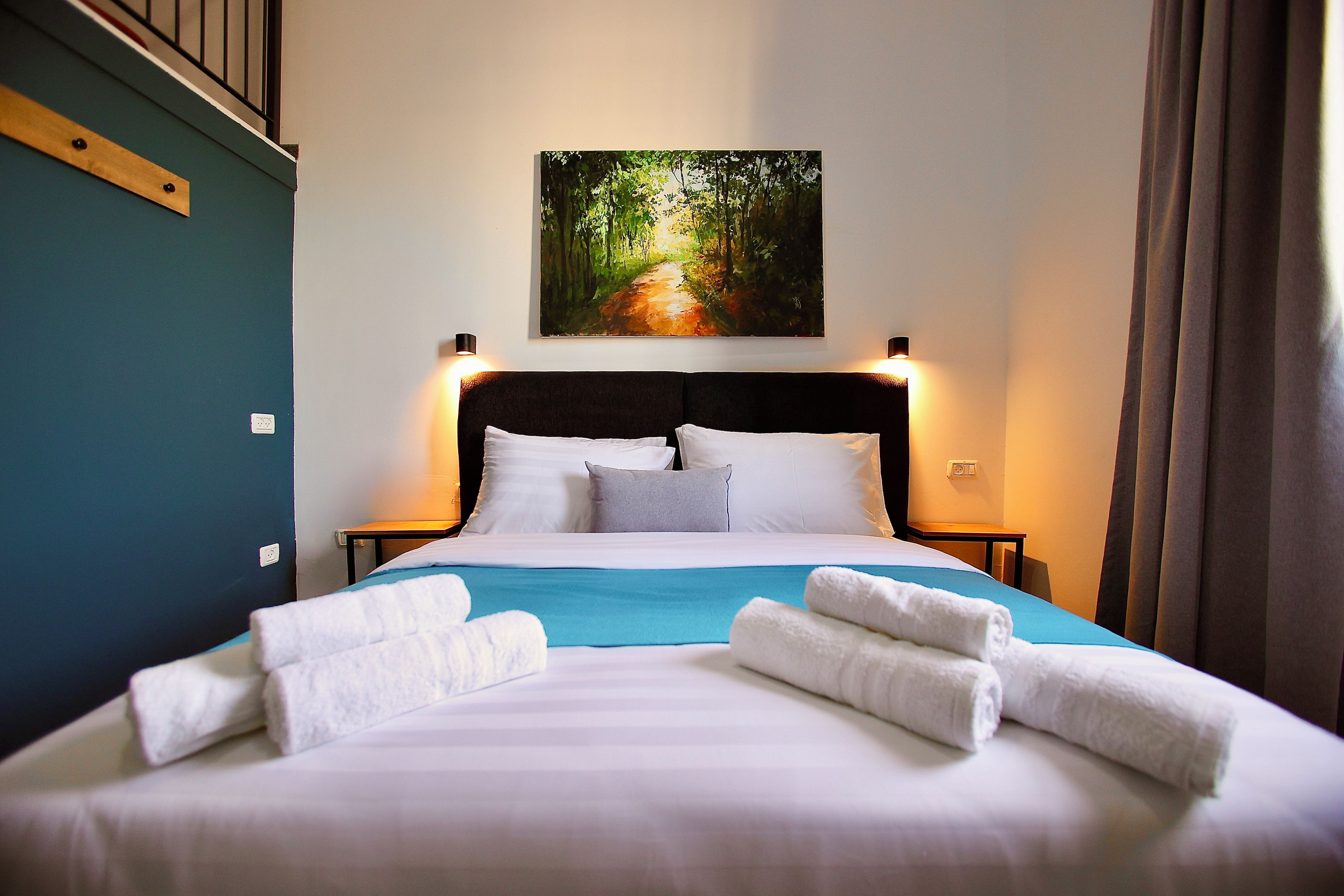 A neatly made bed with white linens and towels, a painting of a forest path above it, and a blue accent wall.
