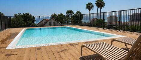 Piscina rectangular con muelle de madera y barandilla metálica, rodeada de palmeras y cielo despejado.