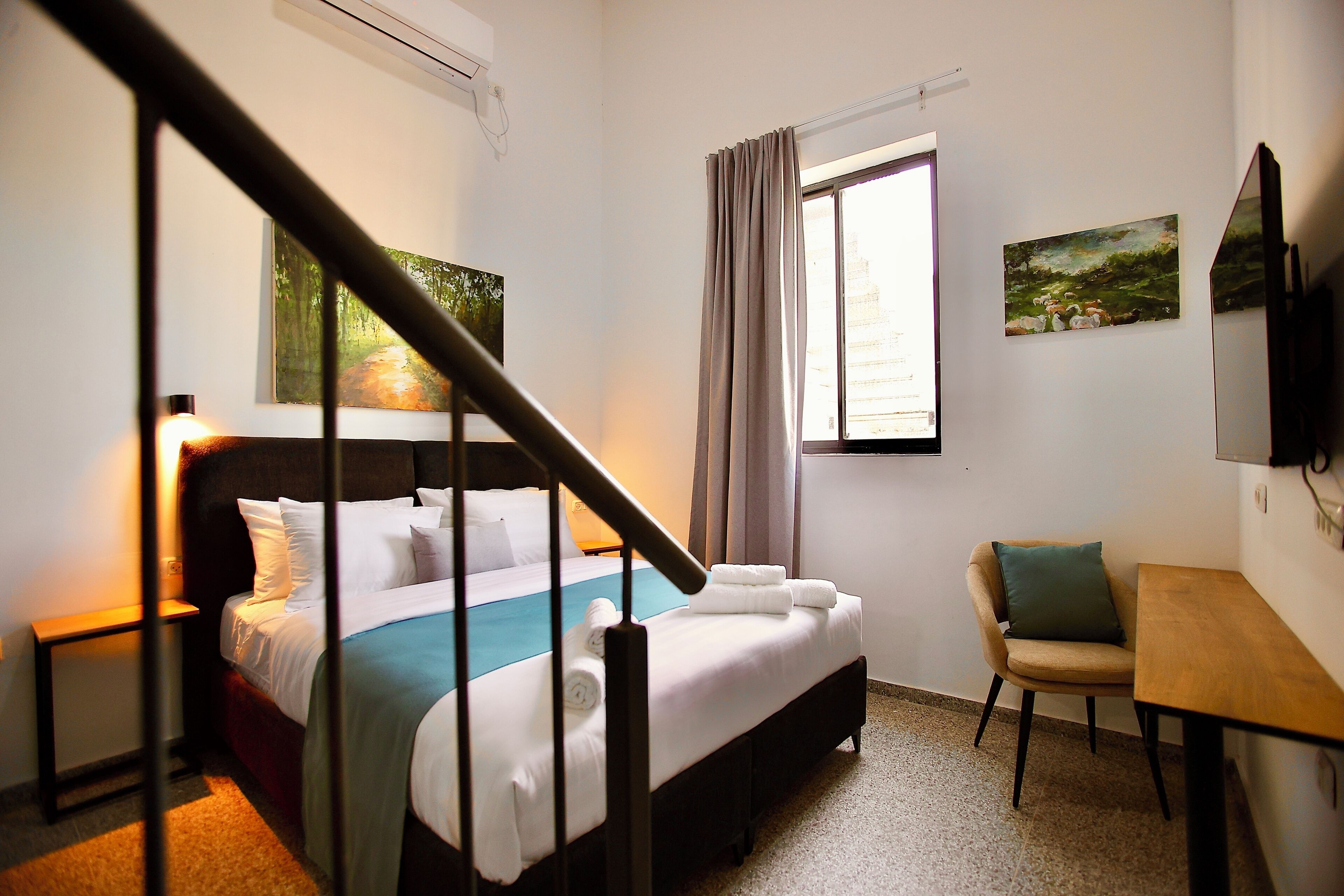 Une chambre d’hôtel comprenant un lit, un bureau, une chaise, une fenêtre avec des rideaux et des œuvres d’art accrochées aux murs.