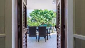 Une porte ouverte donne sur un patio extérieur aménagé avec une table et des chaises, un arbre et une rambarde.