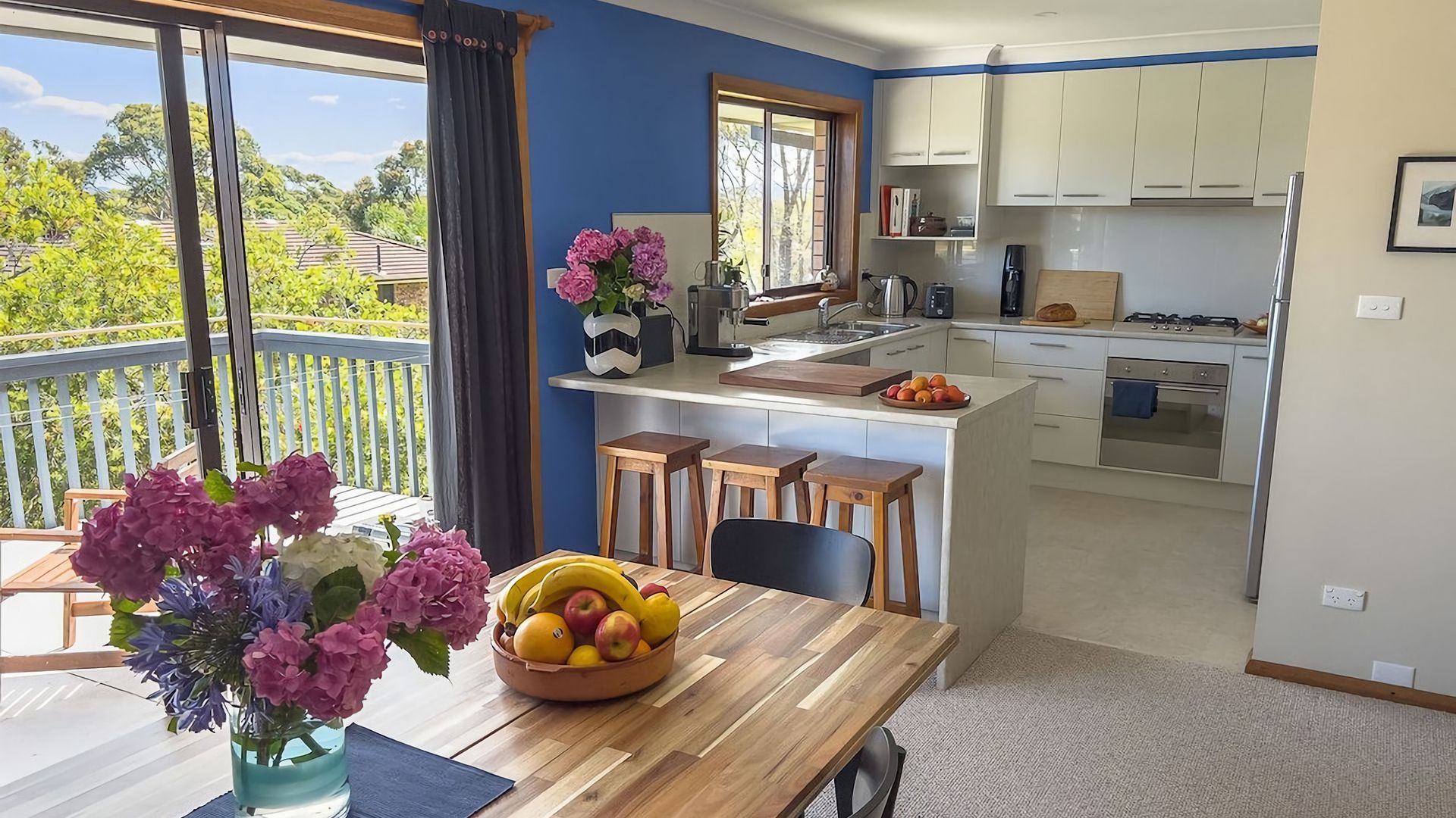 Eine moderne Küche mit weißen Schränken, ein Essbereich mit einem Holztisch und ein Balkon mit Blick auf Grünflächen.
