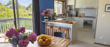 A modern kitchen with white cabinets, a dining area with a wooden table, and a balcony with a view of greenery.