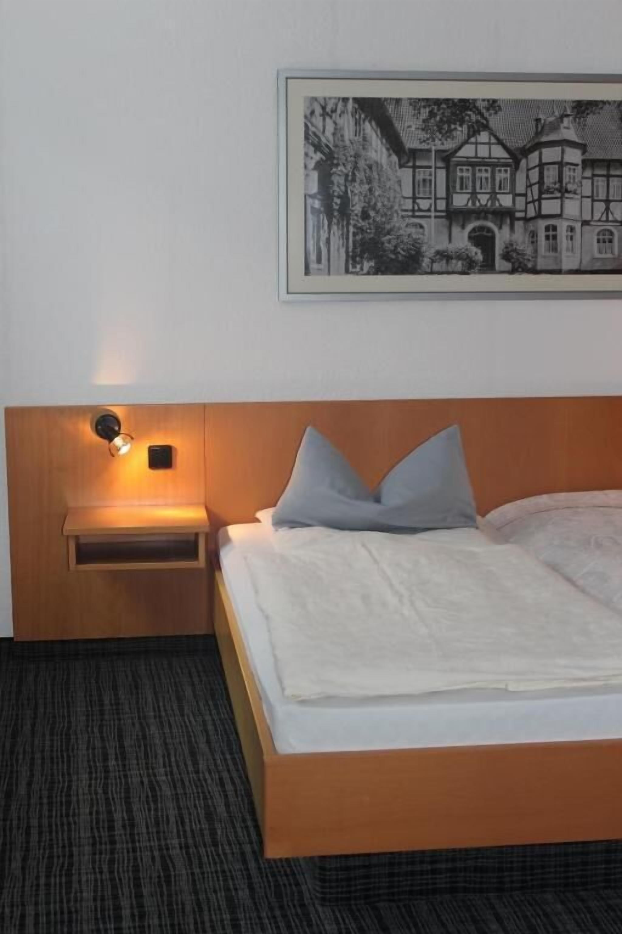 Una camera d'albergo con una testiera in legno, un comodino con una lampada, un letto con lenzuola bianche e una fotografia in bianco e nero incorniciata appesa al muro.