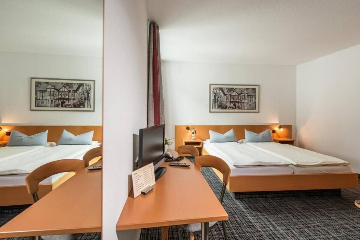 A hotel room with two beds, a desk, a chair, a TV, and framed pictures on the walls.