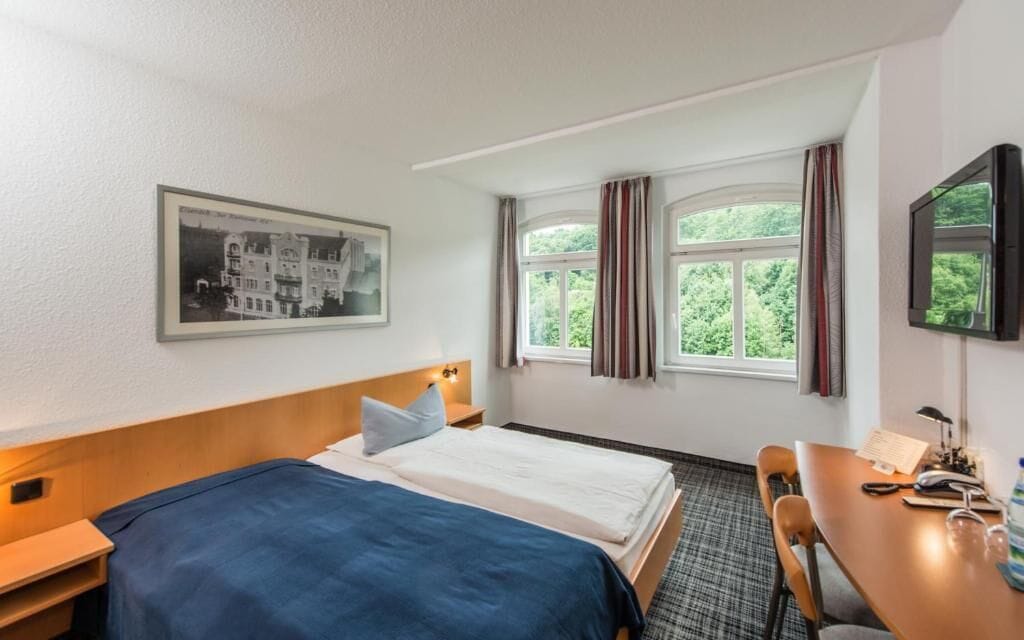 A hotel room with a bed, a desk, a chair, a window with curtains, a framed picture on the wall, and a television.