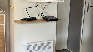 A modern living room with a flat-screen TV mounted on a white cabinet, a small table with a remote control, and a built-in air conditioning unit. - Mobile home in La Palmyre, 10 minutes from the beach, close to the zoo and the Coubre lighthouse (Les Mathes)
