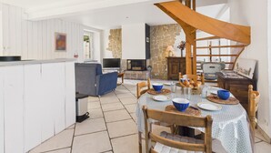 En rummelig opholdsstue med spisebord til fire personer, en stengulvspejs og et højt loft med synlige træbjælker.