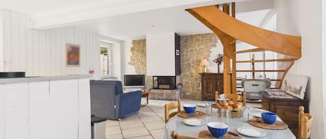 A spacious living area with a dining table set for four, a stone fireplace, and a high ceiling with exposed wooden beams.