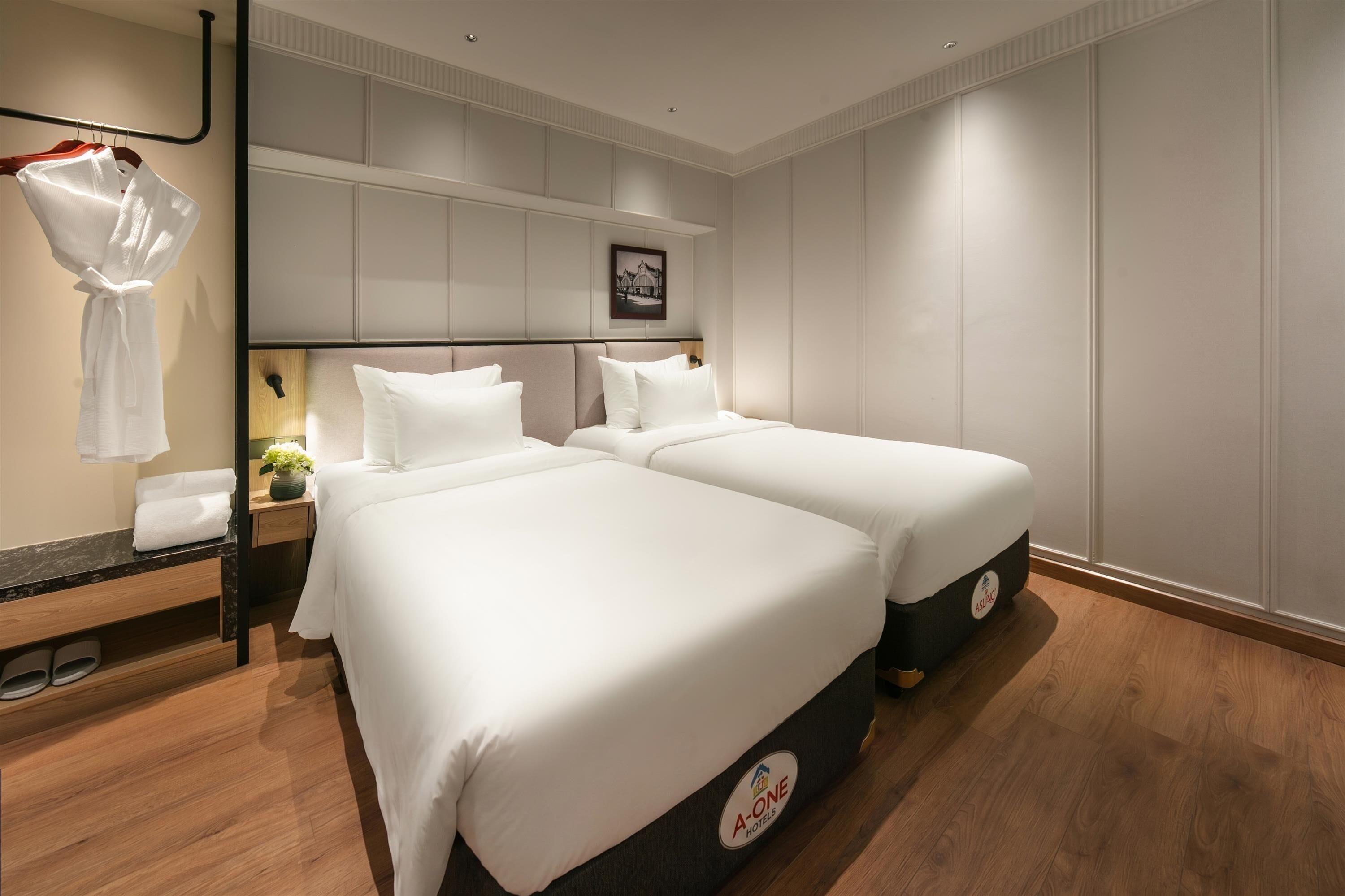 A hotel room with two beds, a nightstand with a lamp, a robe hanging, and a framed picture on the wall.