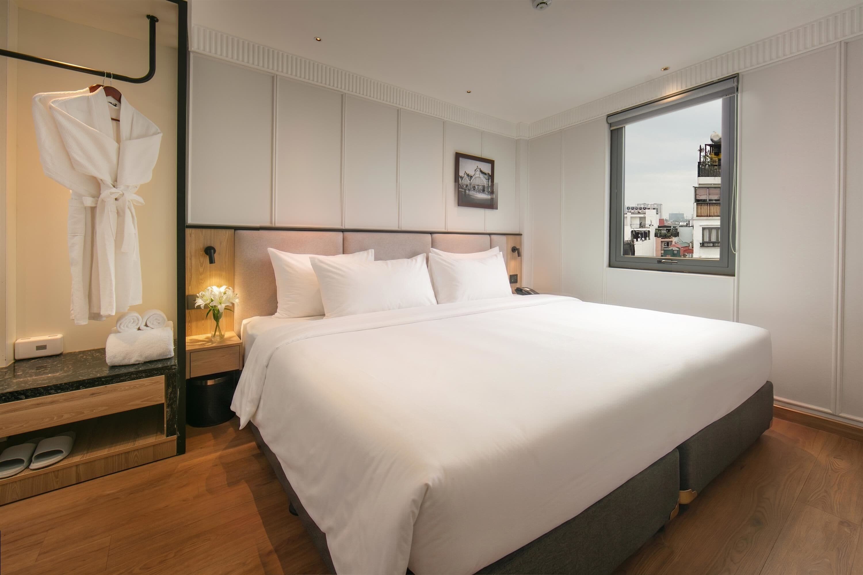 Een hotelkamer met een groot bed, een nachtkastje met een lamp, een spiegel, een badjas hangend aan de muur, en een raam met uitzicht op de stad.