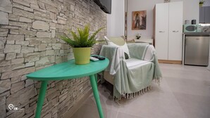 Ein modernes Wohnzimmer mit Steinwand, einem grünen Tisch mit Topfpflanze, einer Couch mit Decke, einem flachbildfernseher über der Couch und einer Küchenzeile mit Mikrowelle und Schränken.
