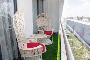 En balkon med to fletstole og et lille bord, gulv af grøn kunstgræs og gelænder af glas med udsigt over bybilledet.