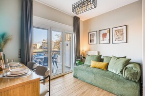 A modern living room with a green sofa, a dining table with plates and glasses, and a balcony with a view of buildings.