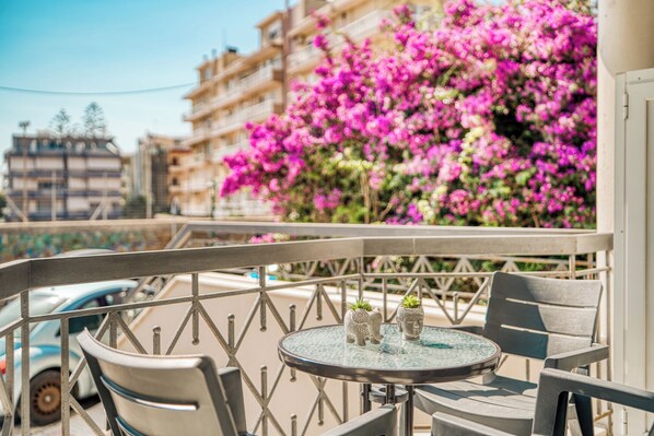 A balcony with a table and chairs, a railing, and a view of pink flowers and buildings.
