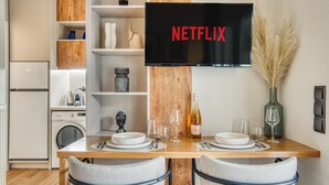 Una cuina moderna amb taula de dinar de fusta per a dues persones, televisió penjada sobre la taula que mostra el logotip de Netflix, i un ram de plomes d'avet en un vas decoratiu.
