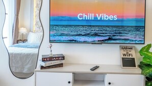 Une chambre d’hôtel moderne avec un grand téléviseur affichant l’inscription « Ambiance détendue », un bureau blanc avec une télécommande, un panneau indiquant « Bienvenue sur notre réseau Wi-Fi » et une petite plante posée sur le bureau.