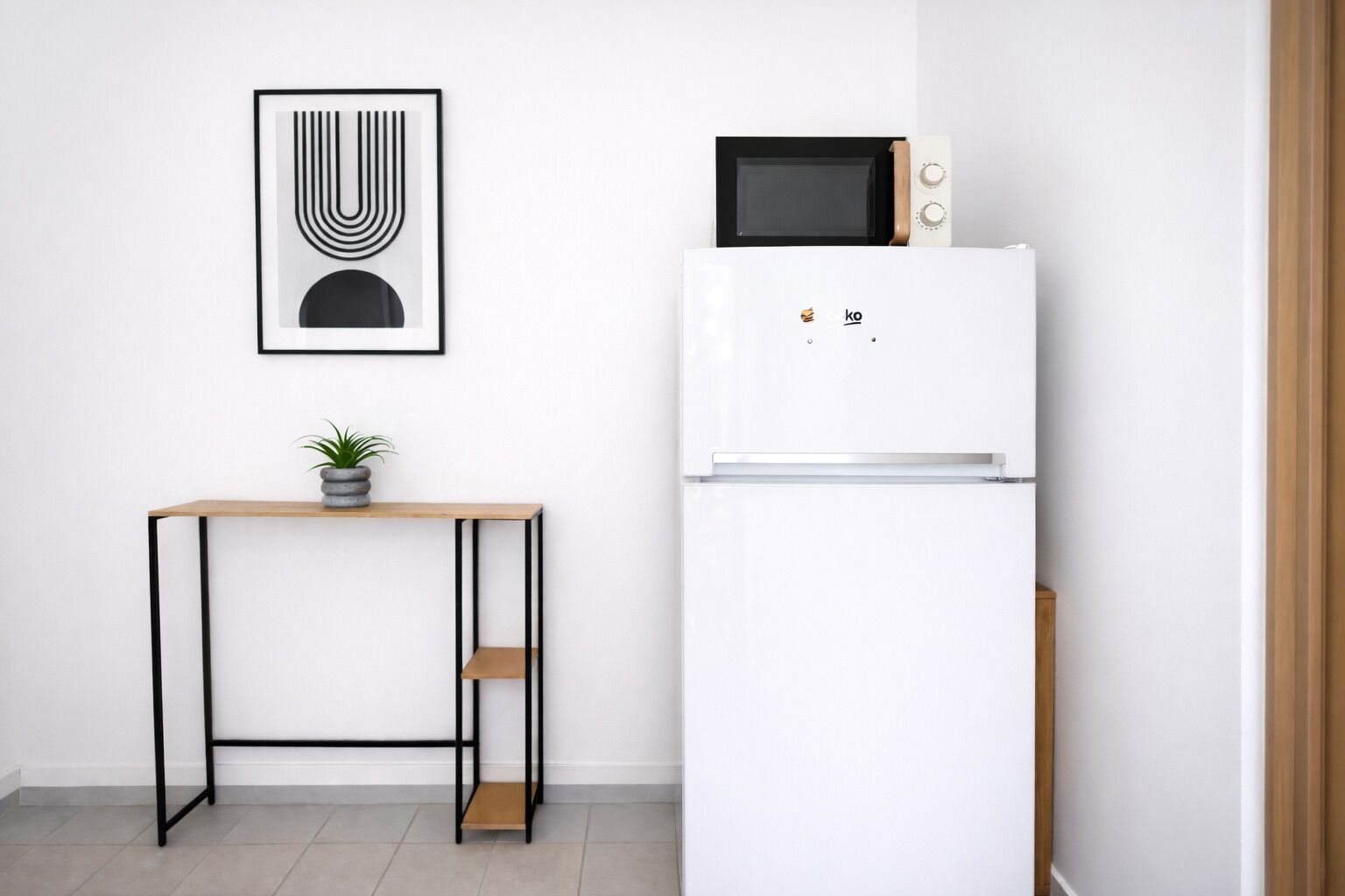 A compact kitchen with a white refrigerator, a microwave, a small wooden table with a potted plant, and a framed abstract artwork on the wall.