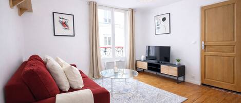 A modern living room with a red sofa, a glass coffee table, a TV on a wooden cabinet, and a large window with curtains.