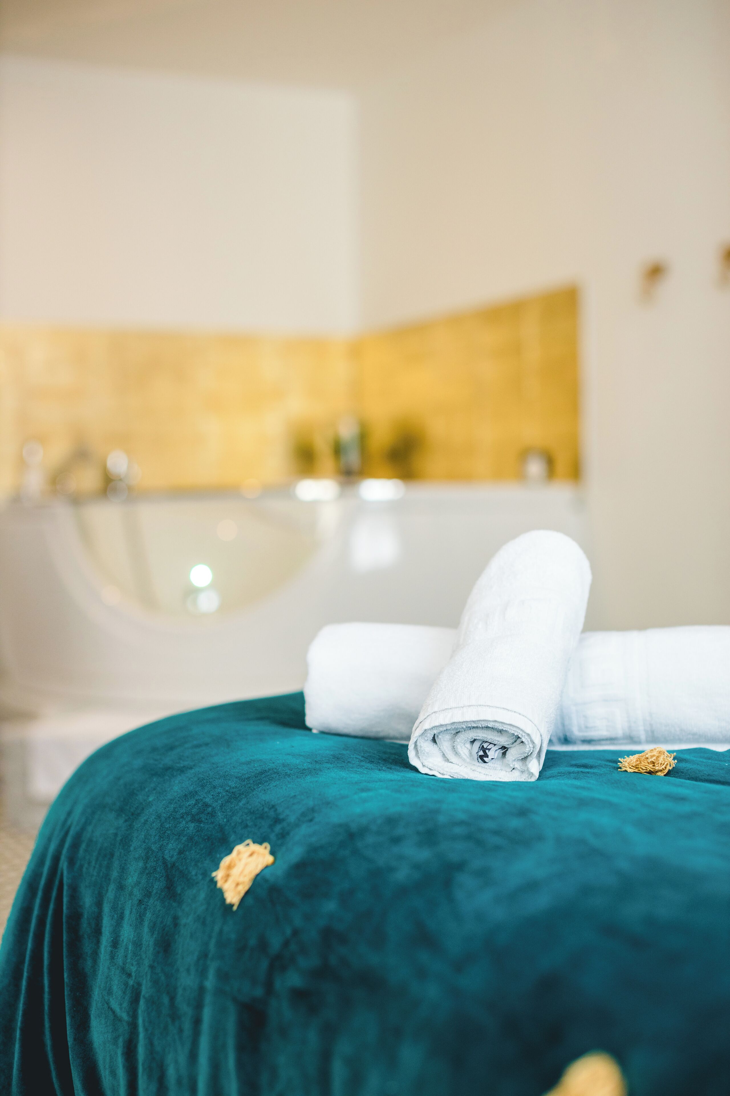 A bed with a teal bedspread and rolled white towels, a bathtub, and a gold-tiled wall in the background.