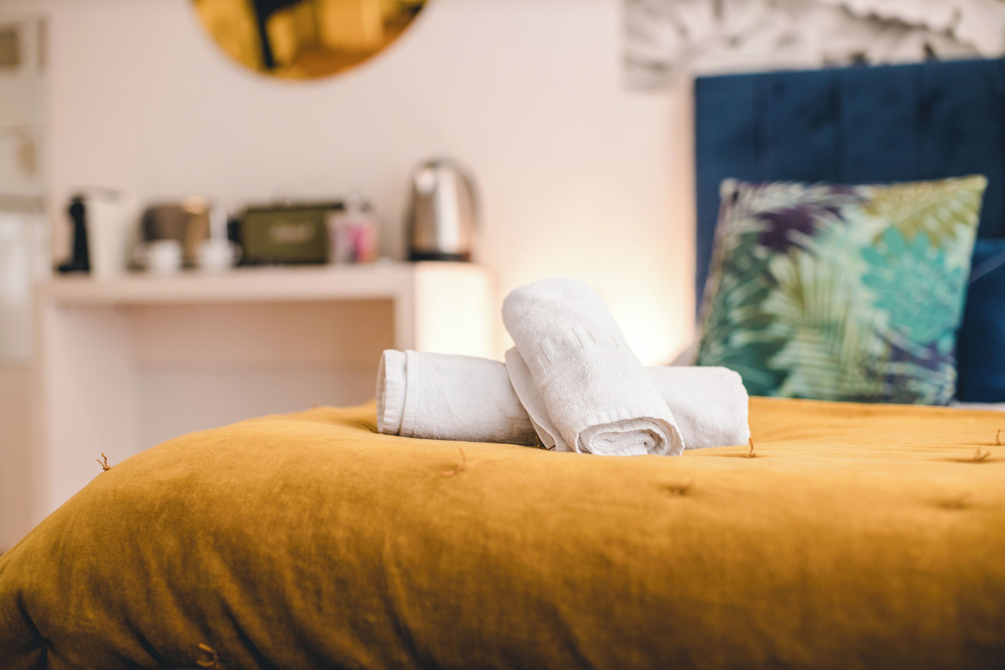 A bed with a mustard-colored duvet, two rolled white towels, a decorative pillow with a tropical leaf pattern, and a white shelf with a kettle and other items in the background.