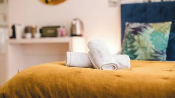 A bed with a mustard-colored duvet, two rolled white towels, a decorative pillow with a tropical leaf pattern, and a white shelf with a kettle and other items in the background.