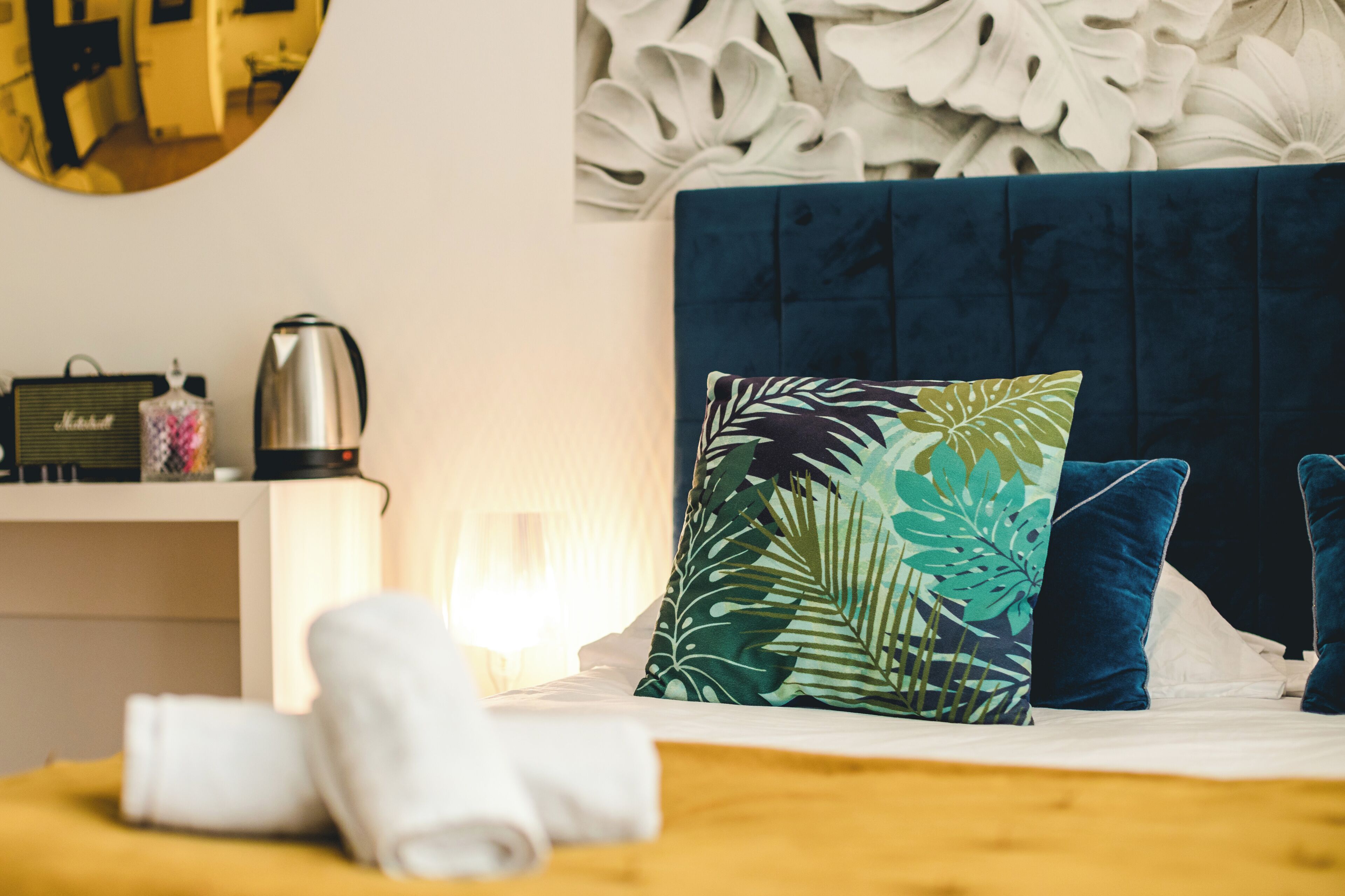 Una cama bien hecha con un cabecero azul, una almohada estampada con motivos de hojas tropicales y una toalla blanca al lado.