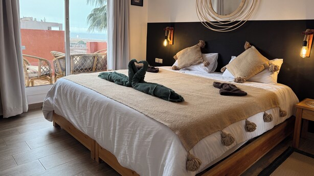 Une chambre d’hôtel avec un grand lit, une vue sur l’océan et un balcon aménagé avec des meubles en osier.