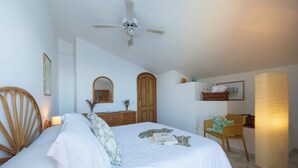 A bedroom with a bed, wooden headboard, a ceiling fan, a bedside table with a lamp, a chair, and a wall-mounted shelf with decorative items. - Sea Whisper Home -Sunset, Sea, Easy Stay, Shops (Praiano)