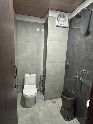 A compact bathroom with a toilet, a shower, and a fan mounted on the wall. - Hotel Utsav (Khatima)