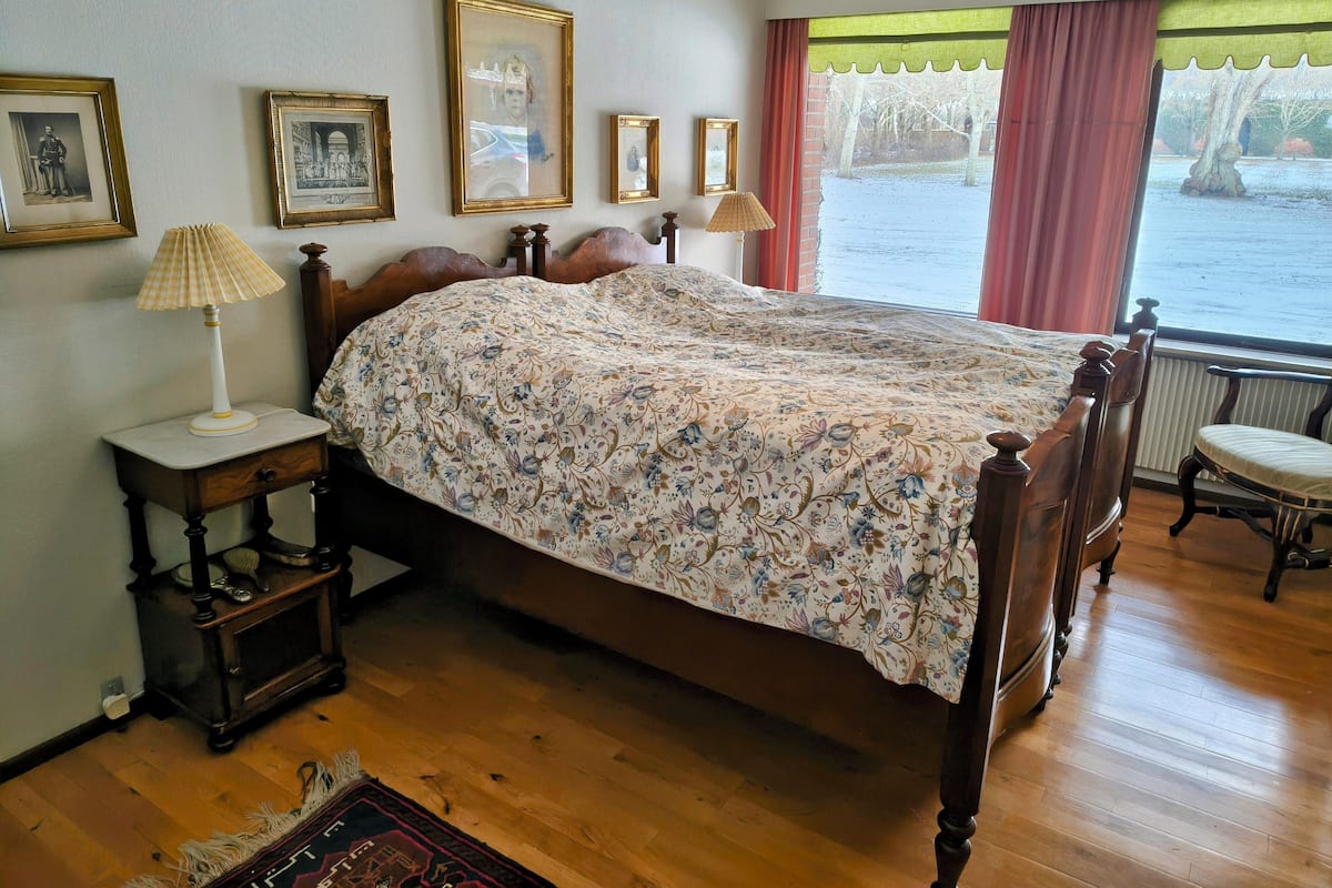 Un dormitorio con cama de madera, una mesita de noche con lámpara, un armario pequeño y vistas a una masa de agua a través de una ventana con cortinas.