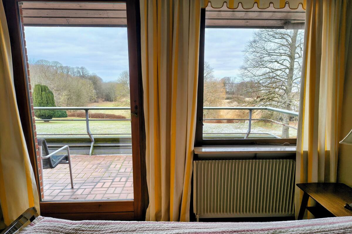 Una habitación con cama, ventana con cortinas, radiador y vistas a un jardín con banco y árboles.