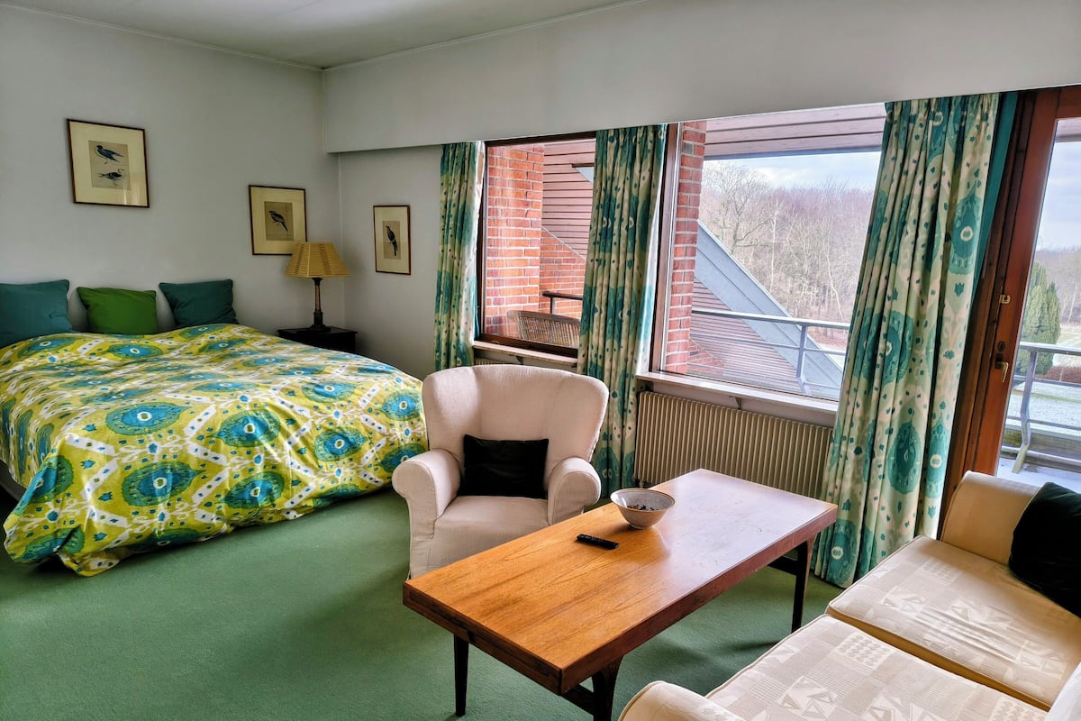 Habitación de hotel con cama, dos sillones, una mesa de madera, un radiador y una ventana con cortinas.