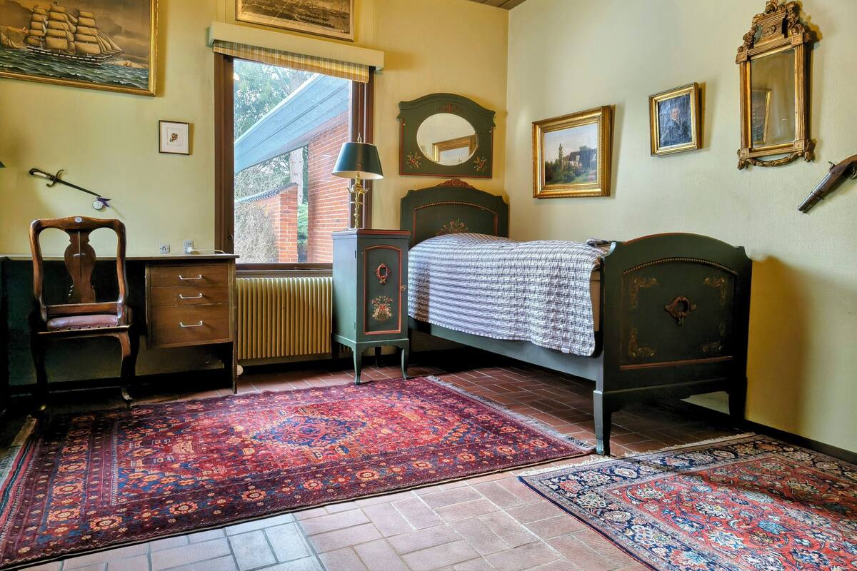 Una habitación con una cama, una silla, un aparador, un radiador y una ventana con vistas a un edificio de ladrillo.