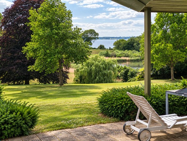 A covered patio with a lawn, a water body in the background, and a lounge chair. - Sæbygaard guesthouse (Hong)