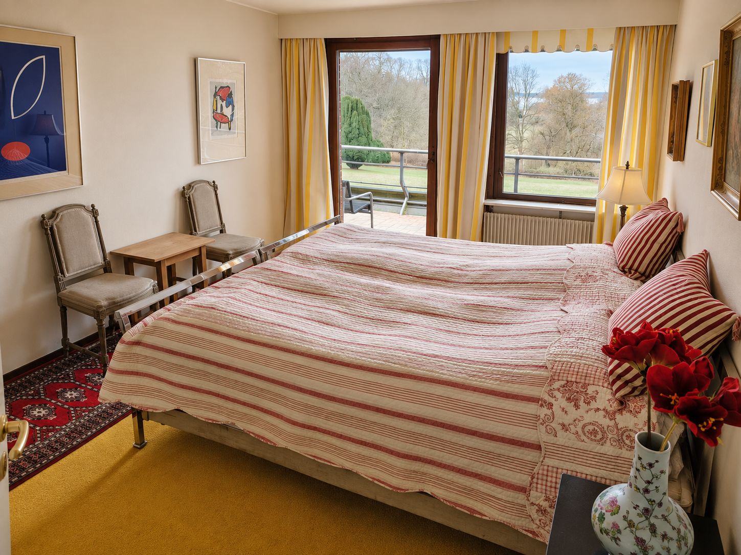 Habitación de hotel con una cama, una silla, una mesita, un florero con flores y vistas al exterior.