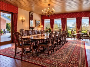 Een eetzaal met een lange tafel, stoelen, een kroonluchter en uitzicht op het groen.