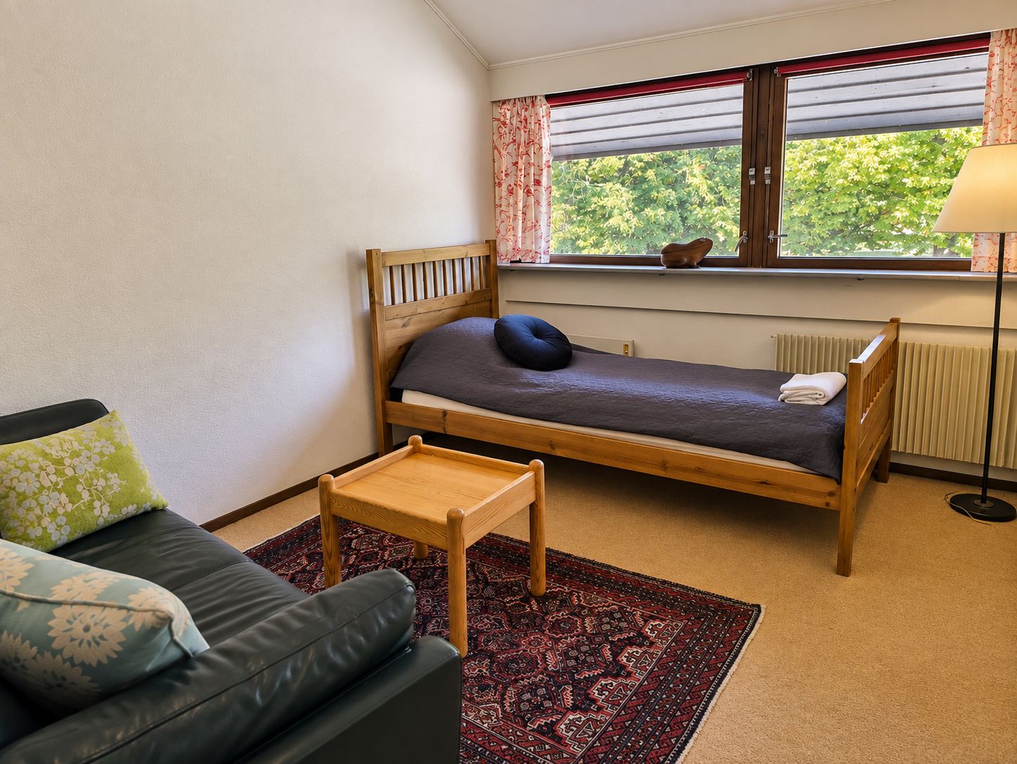 Ein Einzelbett mit Holzrahmen, ein kleiner Nachttisch, ein grünes Sofa mit gemusterten Kissen und ein Fenster mit Vorhängen.