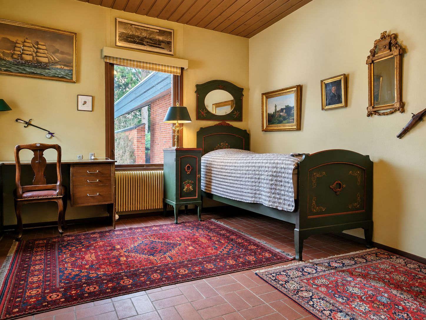 Habitación tradicional con cama, escritorio, silla, radiador y ventana con vista a un edificio de ladrillo.