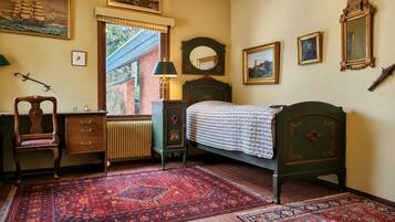 Habitación tradicional con cama, escritorio, silla, radiador y ventana con vista a un edificio de ladrillo.