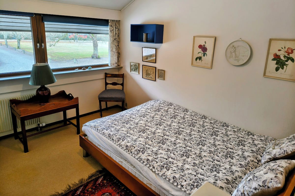 Un dormitorio con cama, una silla, una mesa, un ventanal con cortinas y una obra de arte en la pared.