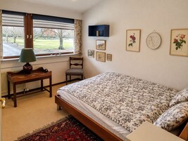 Un dormitorio con cama, una silla, una mesa con lámpara, una ventana con cortinas y cuadros en la pared.