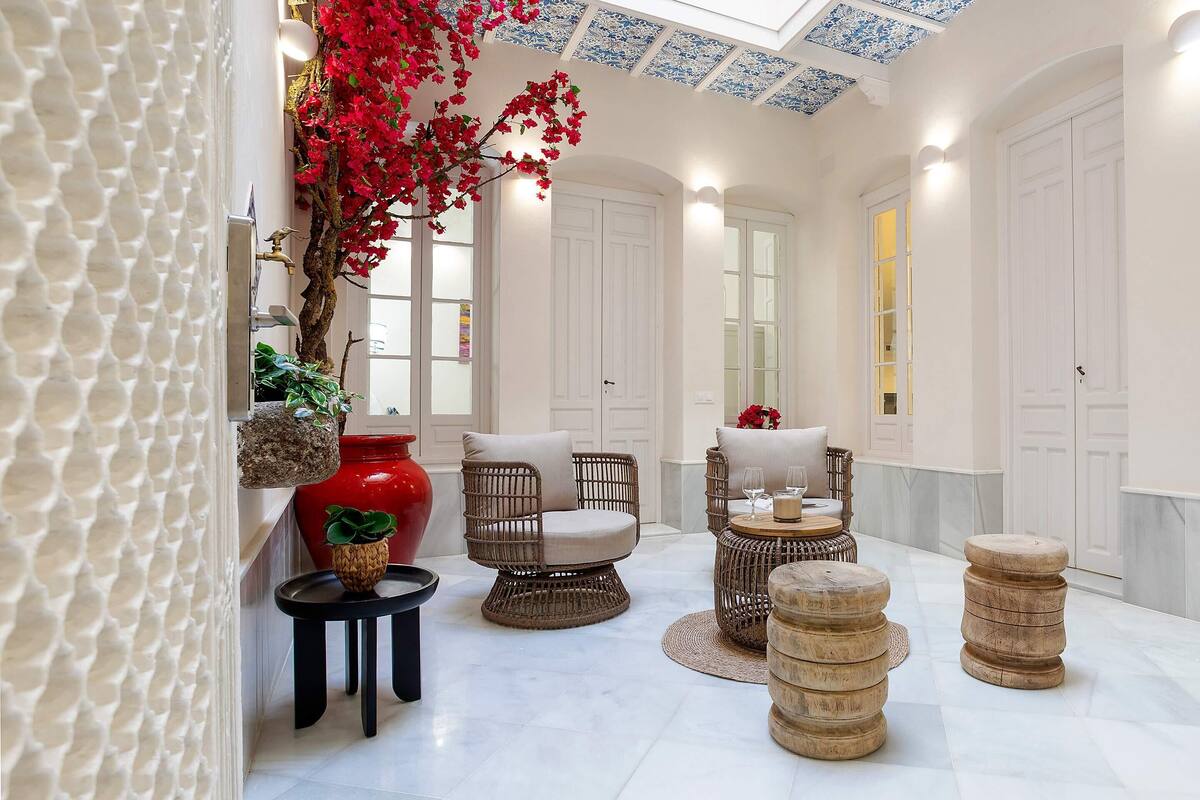 Una habitació àmplia amb un sostre alt, parets blanques i un paviment de rajoles. Hi ha cadires de vimet, una taula rodona petita i un vas vermell amb flors.