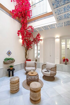 A spacious indoor area with a high ceiling, a large tree with red flowers, wicker furniture, and a tiled floor. - Genteel Home Mendoza Ríos (Seville)