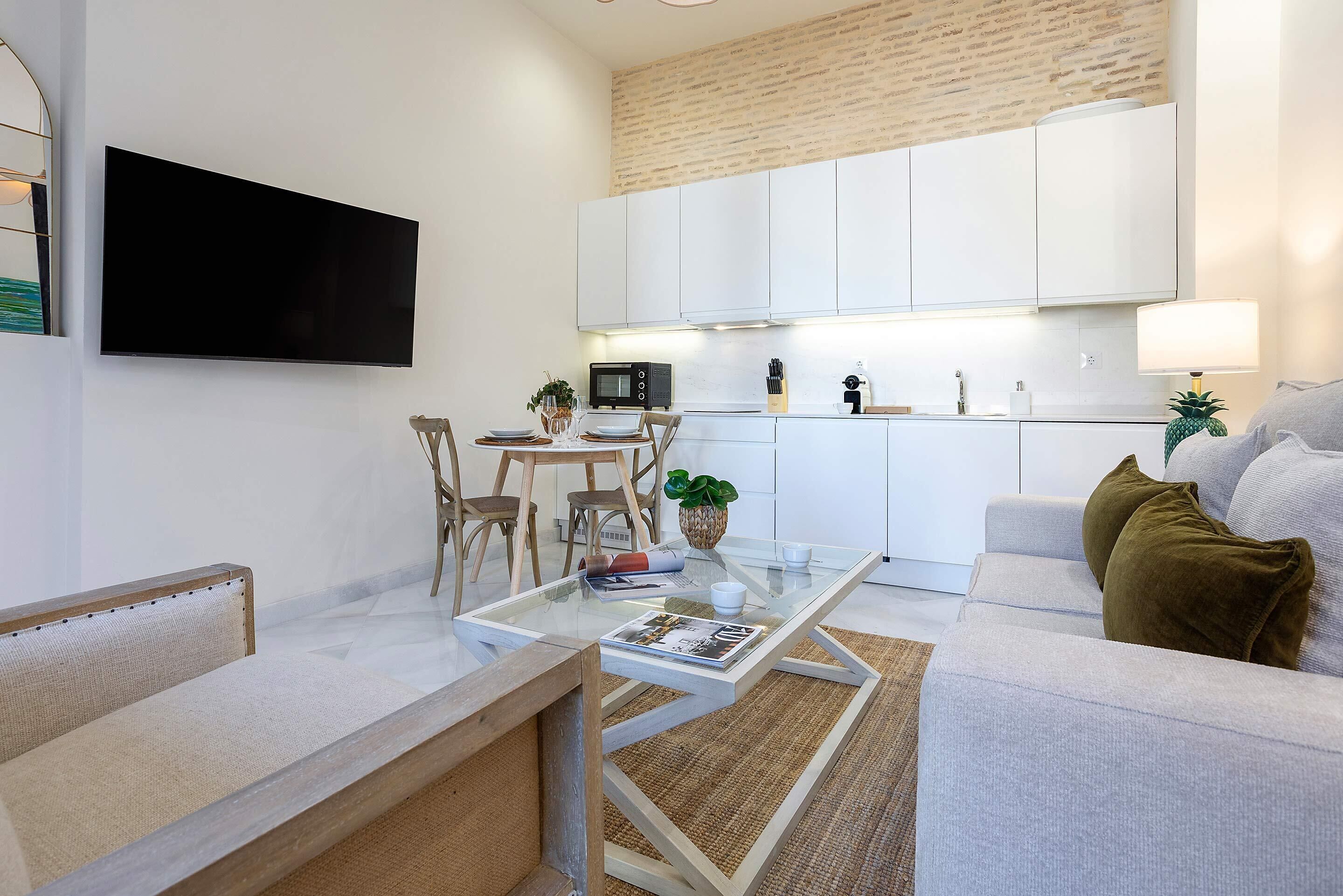 A modern living room with a glass coffee table, a sofa, a TV mounted on the wall, and a kitchen area with white cabinets and a microwave.