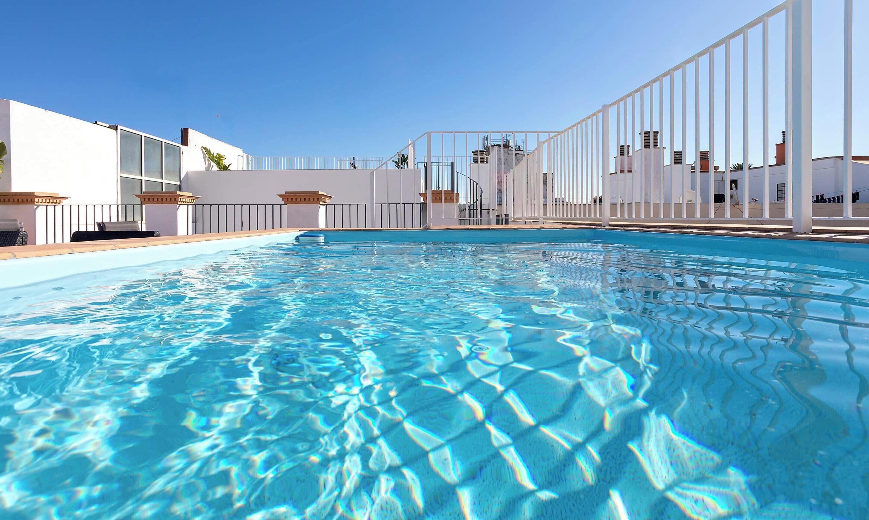 Una piscina con agua azul cristalina, rodeada de muros y una cerca blancos.
