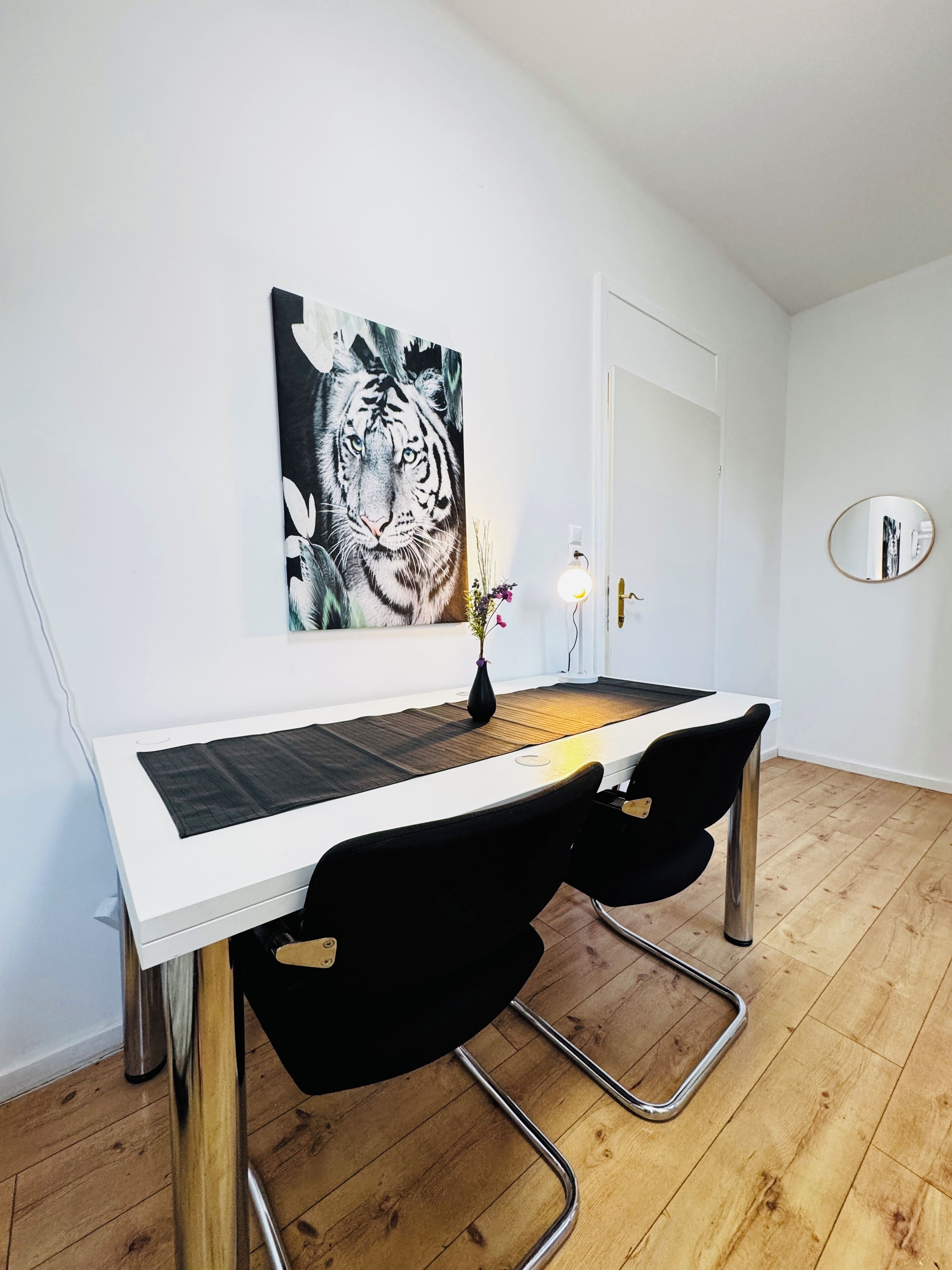 A modern workspace with a white desk, black chairs, a tiger painting, and a round mirror on the wall.
