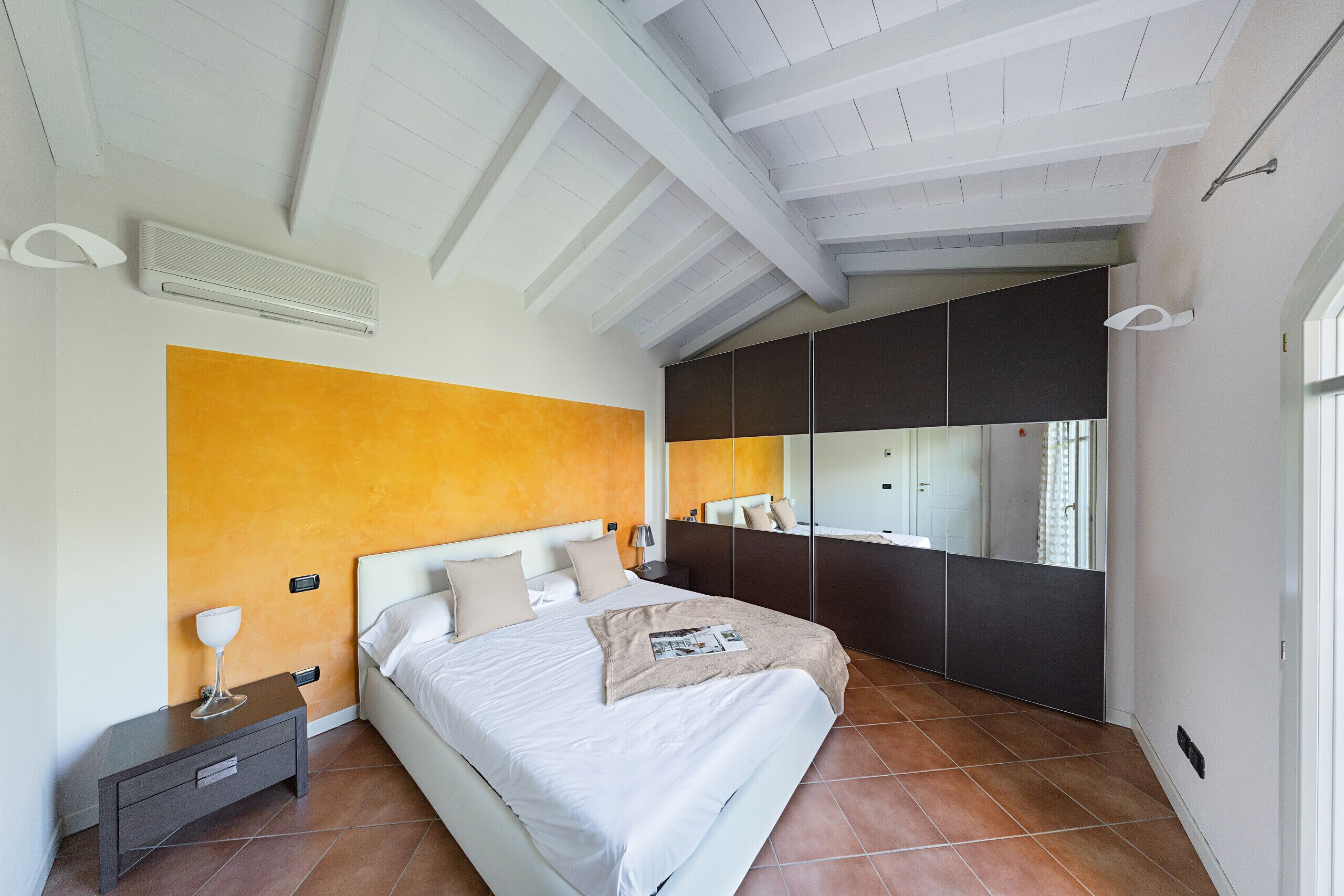 A modern bedroom with a large bed, a nightstand, a bedside table, a mirror, and a window.