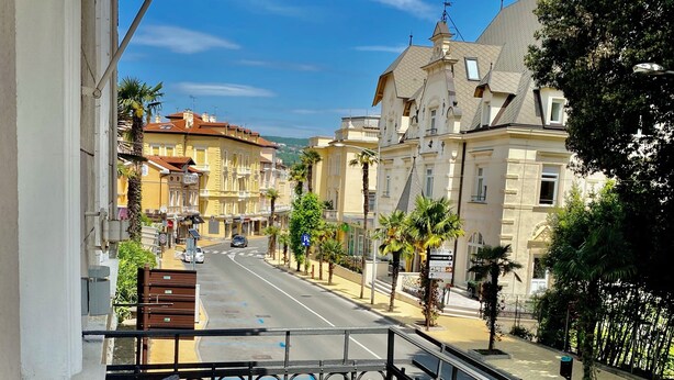 A balcony with a wicker chair and a small table, overlooking a street with buildings and palm trees. - Villa Sokol - Belle Epoque studio (Opatija)